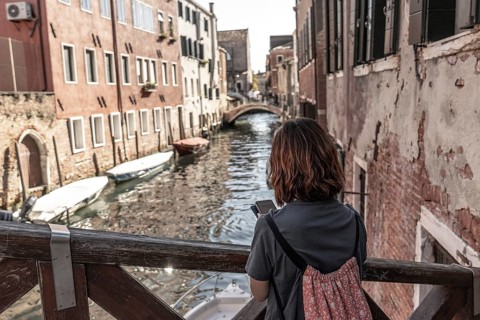 Young woman tourist with backpack using smartphone and enjoying view of picturesque canal, bridge,