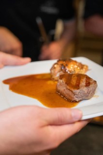 Hand holding a plate with two pieces of roasted meat and plenty of sauce in a kitchen, event and