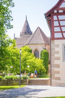 Church tower towers over green trees next to a half-timbered building in summer weather, event and