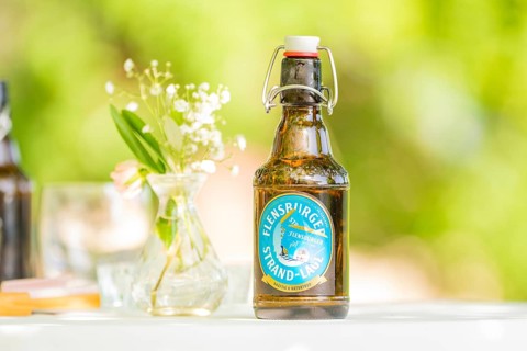 Bottle of Flensburger Strand beer on a table next to a vase of flowers in a green, natural setting,