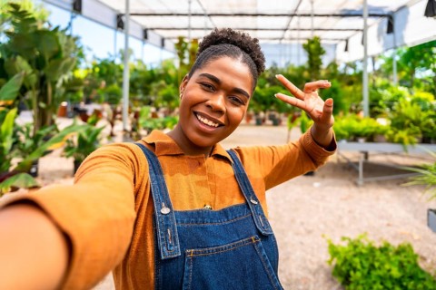 Happy young woman gardener taking a selfie and making a victory sign in a greenhouse full of plants