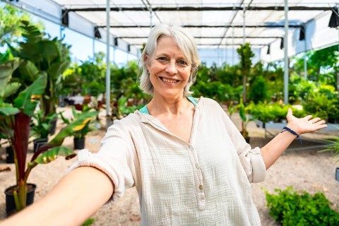 Senior gardener smiling while taking a selfie, proudly displaying vibrant plants in her lush