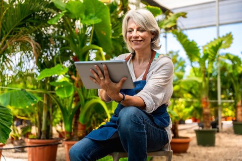 Happy mature woman working in plant nursery and using digital tablet, managing plants and flowers