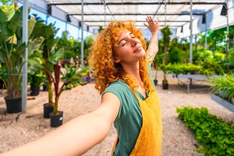 Young woman gardener with curly red hair enjoying the scent of plants, taking a selfie while