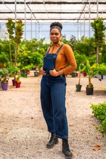 Young african american woman, dressed in denim overalls, posing confidently in a greenhouse,