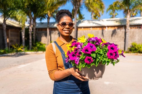 Smiling gardener holding a large pot filled with vibrant, colorful flowers, surrounded by a sunny