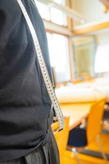 A man in a black T-shirt carries a tape measure over his shoulder in a light-coloured room with