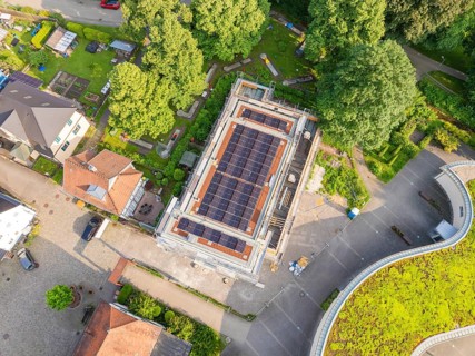 Aerial view of a construction site with solar modules on the roof, surrounded by gardens and