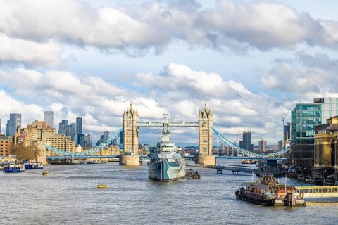 Hms belfast, a town class light cruiser, sailing on the river thames with tower bridge and cloudy
