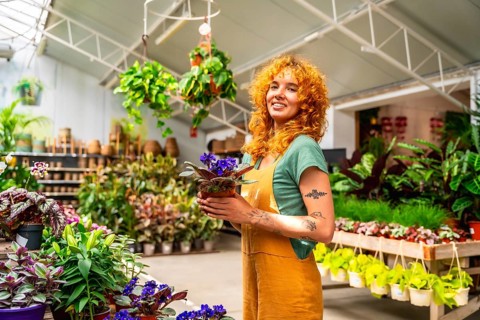Young saleswoman holding a vibrant potted plant and smiling brightly in a lush greenhouse filled