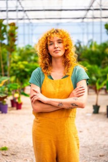 Young woman with curly red hair and tattoos wearing yellow overalls, standing with crossed arms in