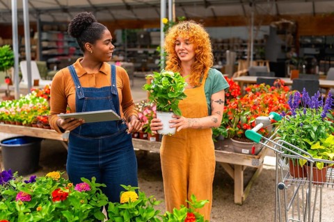 Two young women working in a garden center, discussing plants and using a digital tablet for