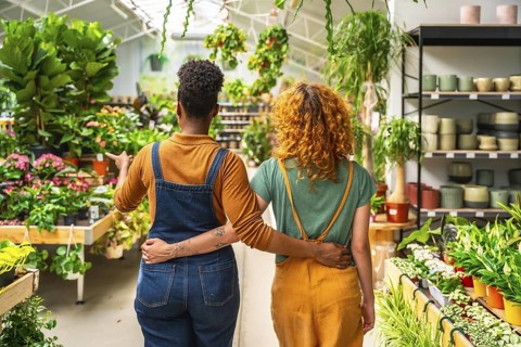 Two young women working in a garden center walking and discussing plants for sale, seen from behind