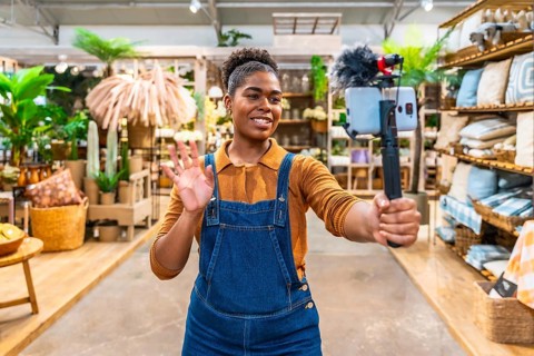 Young saleswoman waving and streaming live video using a smartphone and microphone inside a