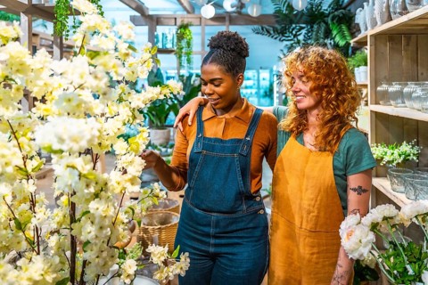 Two smiling female florists are looking and touching white flowers while working together in their