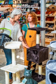 Senior woman holding shopping basket and choosing cat tree with shop assistant help in pet store
