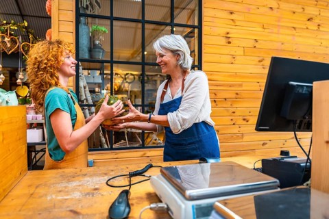 Two women joyfully exchanging a pumpkin in a vibrant flower shop, with a scale and computer