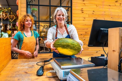 Senior greengrocer carefully weighing a fresh, organic melon in her store, assisted by a younger