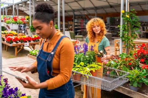 Two young women working in a garden center, taking care of plants and using a digital tablet for