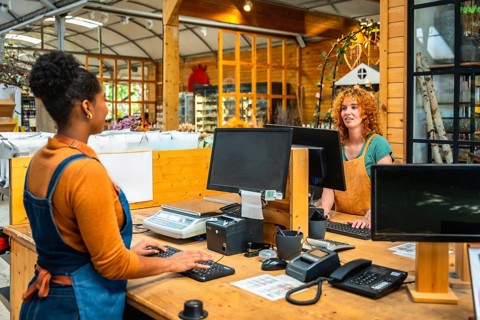 Two young shop assistants are working at the checkout of a plant nursery, using computers and a