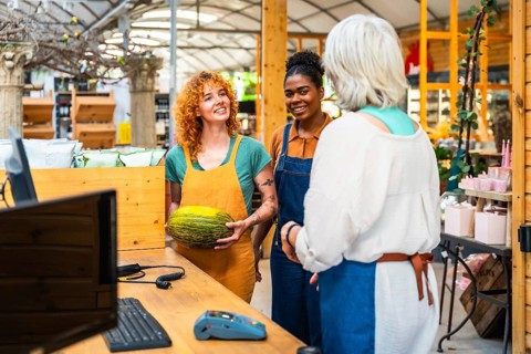 Two young shop assistants engaging with a customer while showcasing a fresh melon in a vibrant