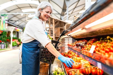 Senior greengrocer with gloves arranging fresh tomatoes on display at farmers market, small