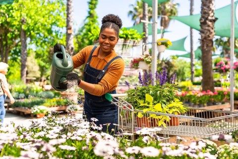 Happy young woman working in a garden center watering plants with a watering can while pushing a