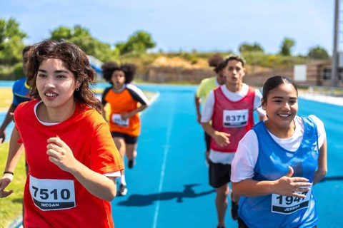 Teenage track and field athletes running on a blue track during a competition, wearing bibs with