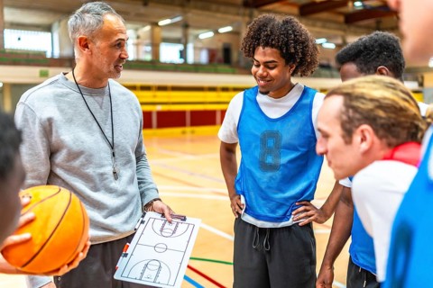 Basketball coach holding ball and clipboard explaining game strategy to team players in gymnasium