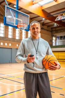 Mature basketball coach holding a ball and stopwatch, smiling confidently in an empty gymnasium,