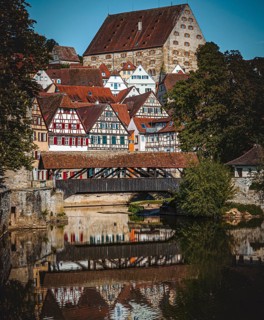 Half-timbered houses with red roofs are reflected in the river next to a wooden bridge in a