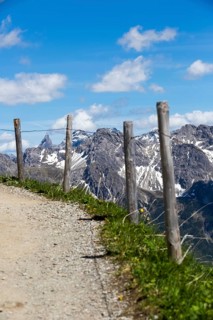 A path lined with wooden fences leads through an alpine landscape with imposing mountain peaks