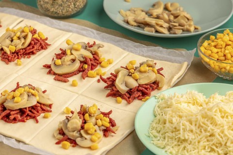 Sliced dough squares with tomato paste, bacon, mushrooms and sweetcorn alongside other ingredients