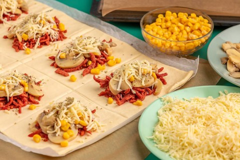 Sliced dough squares with tomato paste, bacon, mushrooms, sweetcorn, cheese and oregano alongside