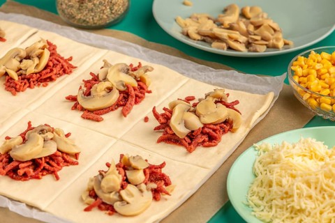 Sliced dough squares with tomato paste, bacon and mushrooms alongside other ingredients for pizza,