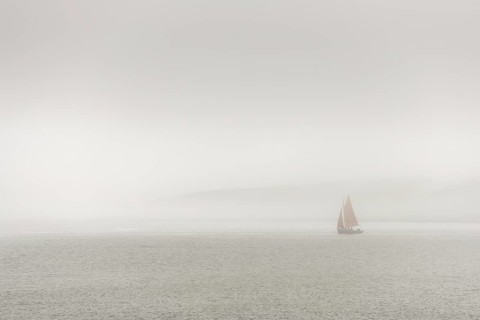 A sailing boat is travelling on the ocean on a cloudy day. The sky is overhanging and the water is