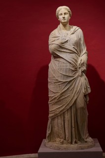 Full-length portrait of an ancient female statue in elaborate drapery against a red background,