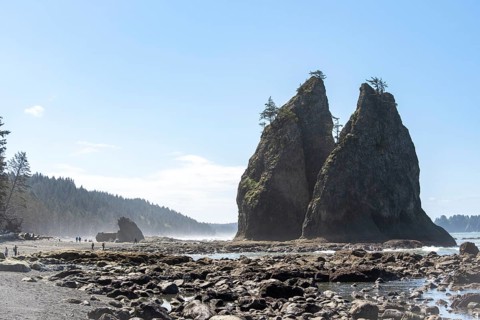 Large rocks protrude from a sandy beach against a clear blue sky, people walk along the shore,