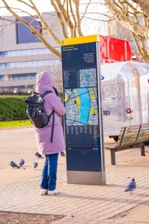 Woman in warm clothes looking at a map at a city map kiosk, pigeons on the ground, London, United