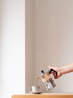 Hand pouring coffee from a moka pot into a cup on a wooden table against a minimalist white