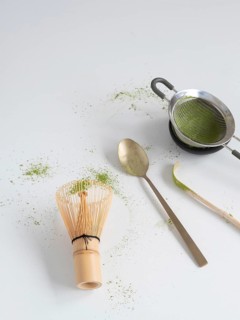 Matcha powder with traditional bamboo whisk, strainer, spoon, and scoop on a white surface