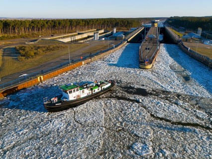 A ship navigates through an ice-covered channel in front of a lock in a wintry landscape with