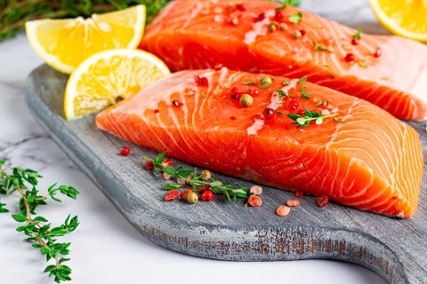 Two pieces of salmon fillet sit on a wooden cutting board. They are topped with herbs and pink