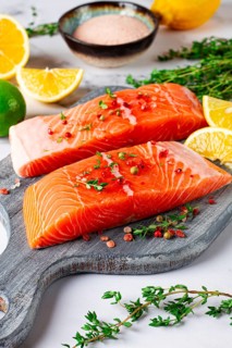Two pieces of salmon fillet sit on a wooden cutting board. They are topped with herbs and pink