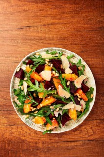 A bowl of salad contains beetroot, orange slices, pine nuts, and leaves. It is placed on a wooden