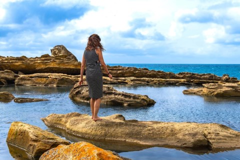 Woman standing barefoot on textured sandstone by tidal pools, facing the vast sea under a cloudy