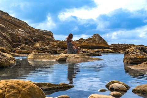 Woman sitting on a rock formation in a coastal landscape, overlooking a tranquil tidal pool,