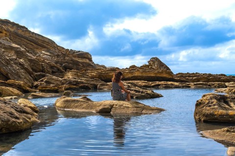 Woman sitting on a rock in a calm tide pool. Reflecting on the water. Surrounded by rugged coastal