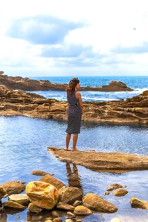 Woman standing barefoot on a rock formation, observing the ocean and the rugged coastline with
