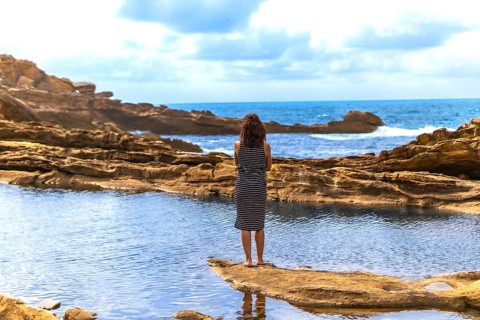 Woman standing barefoot on a rocky outcrop, observing the vast sea and dramatic sky, finding a